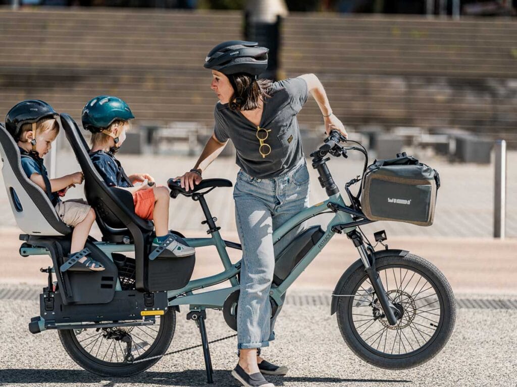 En kvinna som bär hjälm står bredvid hennes Yuba FastRack longtail lastcykel, som är utrustad med en frontmonterad väska. Två hjälmförsedda barn är på cykeln; en sitter i en barnstol medan den andra sitter på den bakre hyllan. De njuter av en solig utomhusmiljö.