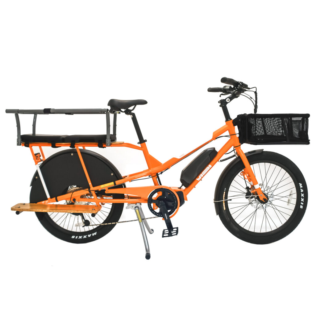 Profilvy av en orange Yuba Kombi E5 Family longtail elektrisk lastcykel med ett bakre passagerarsäte med fotstöd, stort bakhjulsskydd, rymlig framkorg och ett svart rammonterat batteri, visad mot en vit bakgrund.