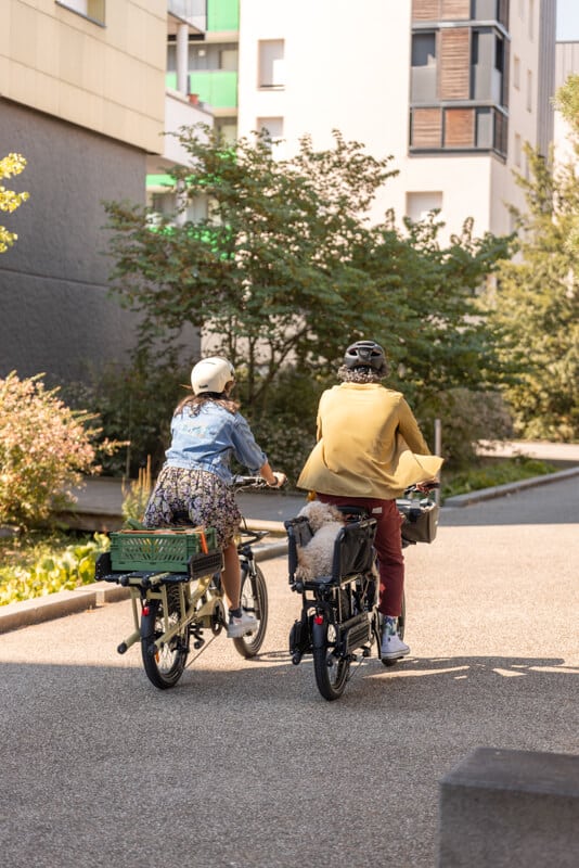 Två personer med hjälm cyklar på Yuba-cyklar längs en solbelyst stig mellan moderna byggnader, omgivna av grönska. Den ena personen cyklar på en Yuba FastRack och bär kjol, medan den andra har röda byxor och en beige jacka och sitter på en annan Yuba-modell. Båda har väskor eller korgar fästa på sina cyklar.