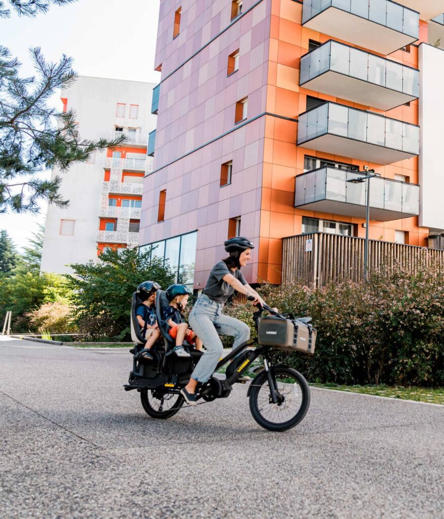 En person cyklar på en Yuba FastRack-lastcykel med två barn sittande bakpå, alla med hjälm på sig, och passerar moderna orange- och vitmålade flerbostadshus och grönområden en solig dag.