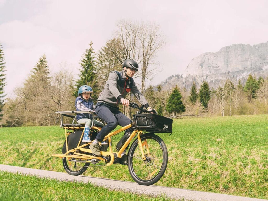 En person som bär hjälm cyklar på en livlig gul Yuba Mundo EP8 Family lastcykel med ett barn som också bär hjälm, sittande glatt i det bakre lastutrymmet. Bakgrunden har frodiga gräsfält, lummiga träd och majestätiska berg under en mulen himmel.