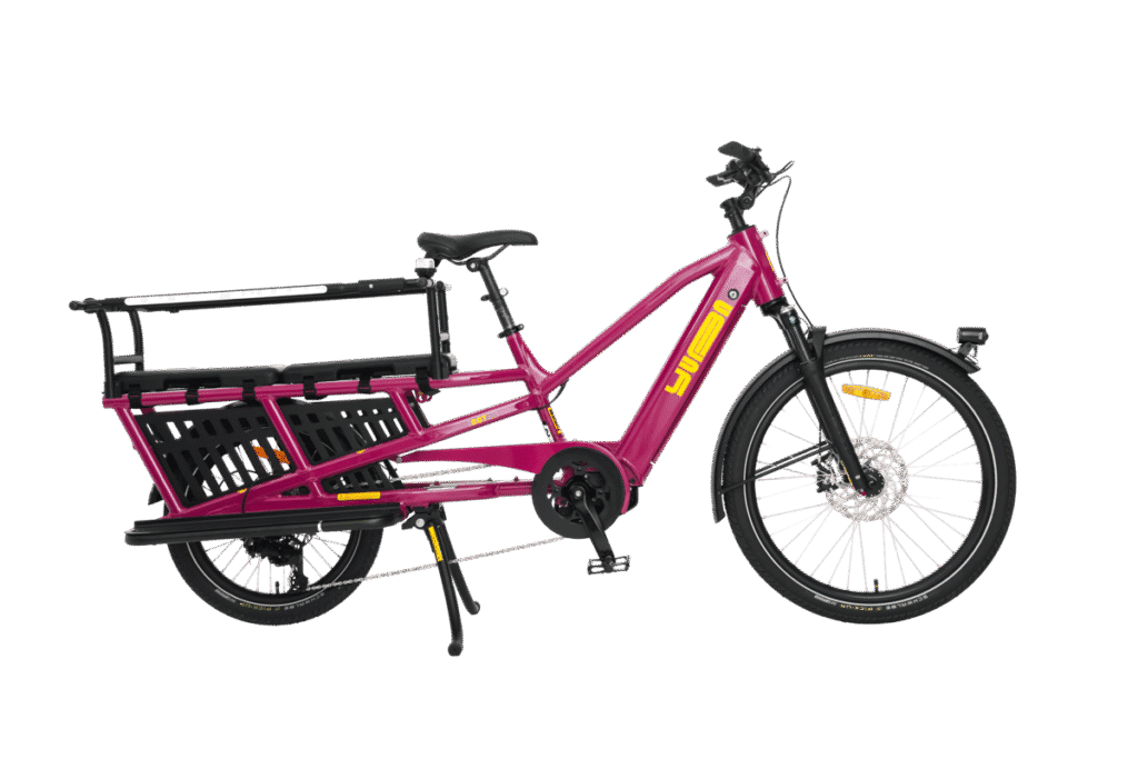 En knallrosa eldriven lastcykel av modellen Yuba Spicy Curry Family Edition med bakre pakethållare, sidokorgar, svart sadel och gula logotyper, avbildad från sidan mot en vit bakgrund. Denna eldrivna longtail-cykel förenar på ett perfekt sätt en livfull stil med praktisk användbarhet i vardagen.