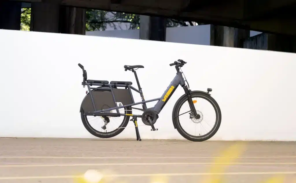 En modern svart och gul Boda Boda longtail elektrisk lastcykel med en robust bakre ställning är parkerad på en asfalterad yta mot en vanlig vit vägg, vilket framhäver dess eleganta design och praktiska möjligheter att bära laster.