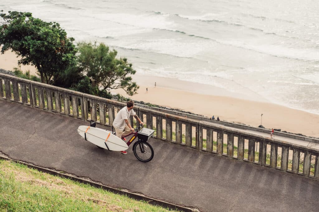 En person i shorts går med en Yuba Spicy Curry Family Edition, en elastisk longtailcykel, med en surfbräda fäst på den, längs en asfalterad stig ovanför en strand, där man kan se havsvågor och några människor nedanför.