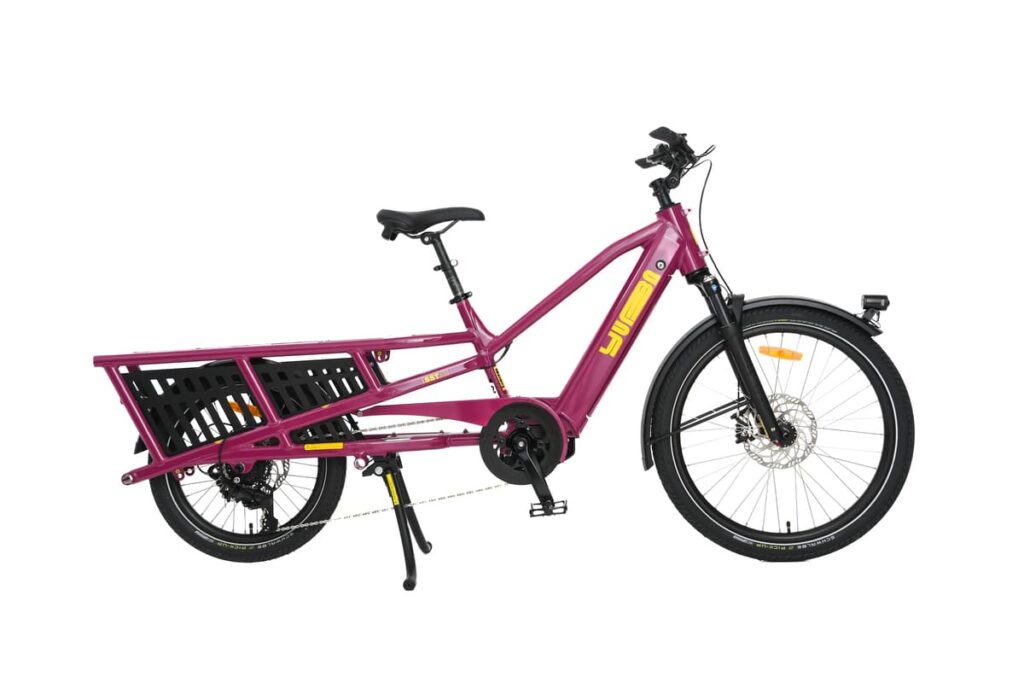 En magenta Yuba Spicy Curry Family Edition, en eldriven lastcykel med ett robust bakre pakethållare, svart korg, stänkskärmar fram och bak samt gula detaljer, avbildas från sidan mot en vit bakgrund. Denna eldrivna longtail-cykel förenar stil och praktiskhet för stadstrafik.