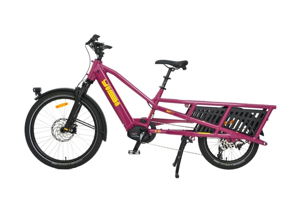 På bilden syns en lila eldriven lastcykel av modellen Yuba Spicy Curry Family Edition mot en vit bakgrund. Den har en robust ram, gul Yuba-logotyp, framfjädring, bakre lastställ med sidoräcken samt ett stöd. Denna eldrivna longtail-cykel är utrustad med en kraftfull Bosch-motor som garanterar pålitlig prestanda och smidiga åkturer.