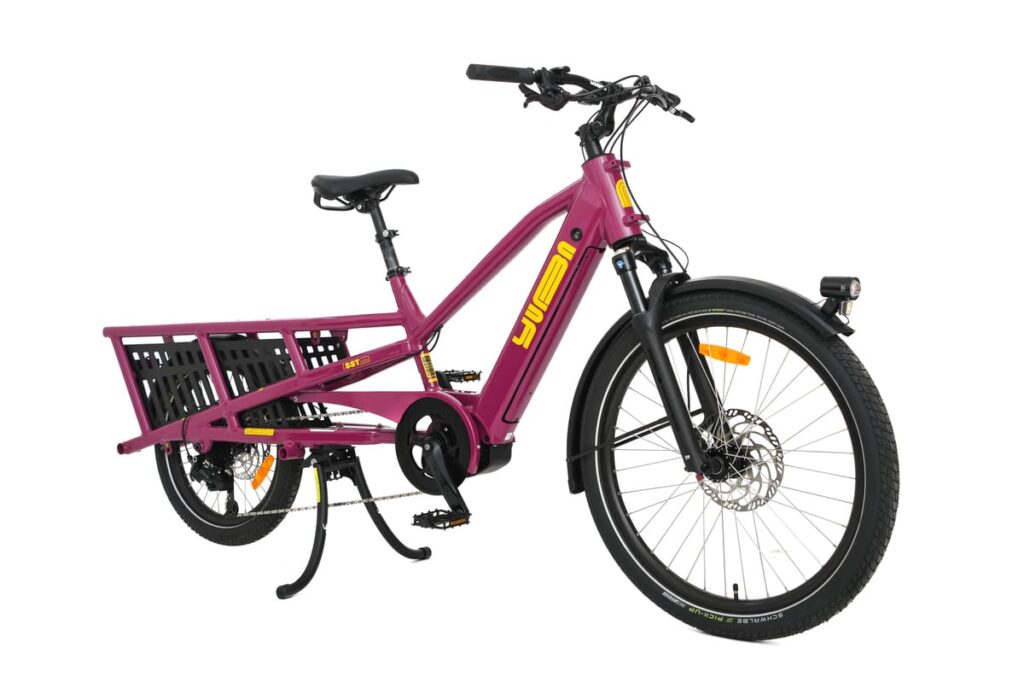 En magentaröd eldriven lastcykel av modellen Yuba Spicy Curry Family Edition, utrustad med ett robust bakre pakethållare, en svart korg, gula dekaler, framfjädring, skivbromsar och ett stöd, är fotograferad mot en vit bakgrund. Denna longtail-modell är utrustad med en kraftfull Bosch-motor som gör det enkelt att transportera last och ger en smidig åktur.