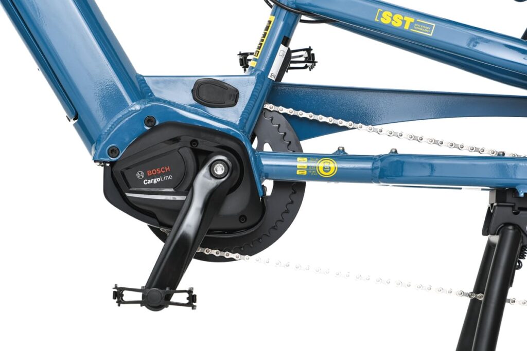 Närbild på den blå cykelramen av modellen Yuba Spicy Curry Family Edition, utrustad med elmotorn Bosch Cargo Line, vevpartiet och kedjan. Bilden visar komponenterna i en elassiterad longtail-cykel och belyser den robusta konstruktionen och kraften hos denna moderna longtail-cykel med Bosch-motor.