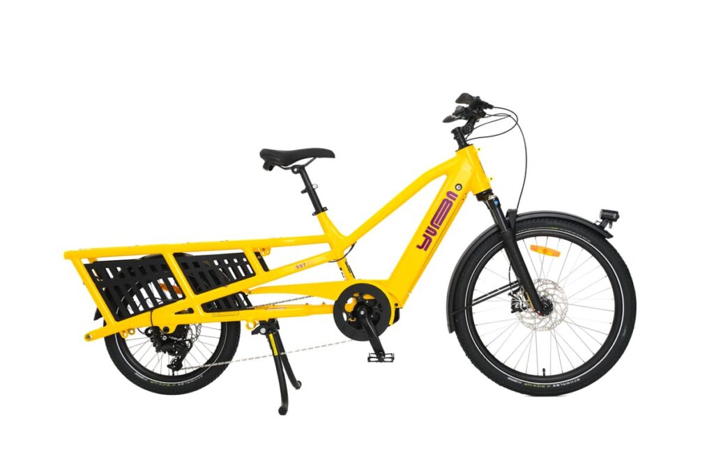 En knallgul Yuba Spicy Curry Family Edition-longtailcykel med ett långt bakre pakethållare, svart sadel, skärmar fram och bak samt ett stöd, fotograferad mot en vit bakgrund.