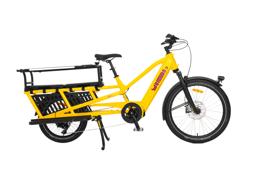 Yuba Spicy Curry Family Edition är en knallgul eldriven lastcykel utrustad med ett robust bakre pakethållare, svart sadel och svart styre, utformad för att transportera passagerare eller last. Denna longtail-modell drivs av en kraftfull Bosch-motor som ger smidiga och bekväma åkturer, och har varumärket ”Yuba” på ramen.