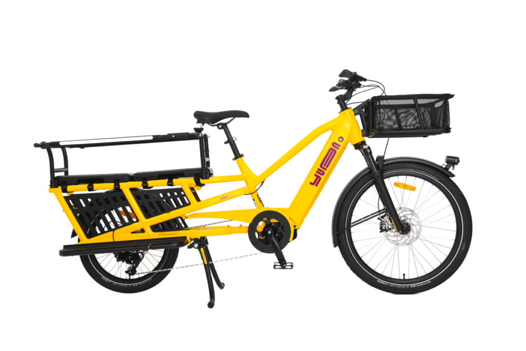 En knallgul Yuba Spicy Curry Family Edition-longtailcykel med Bosch-motor, utrustad med en framkorg, ett bakre passagerarställ och två stora svarta sidokorgar, avbildad mot en vit bakgrund.
