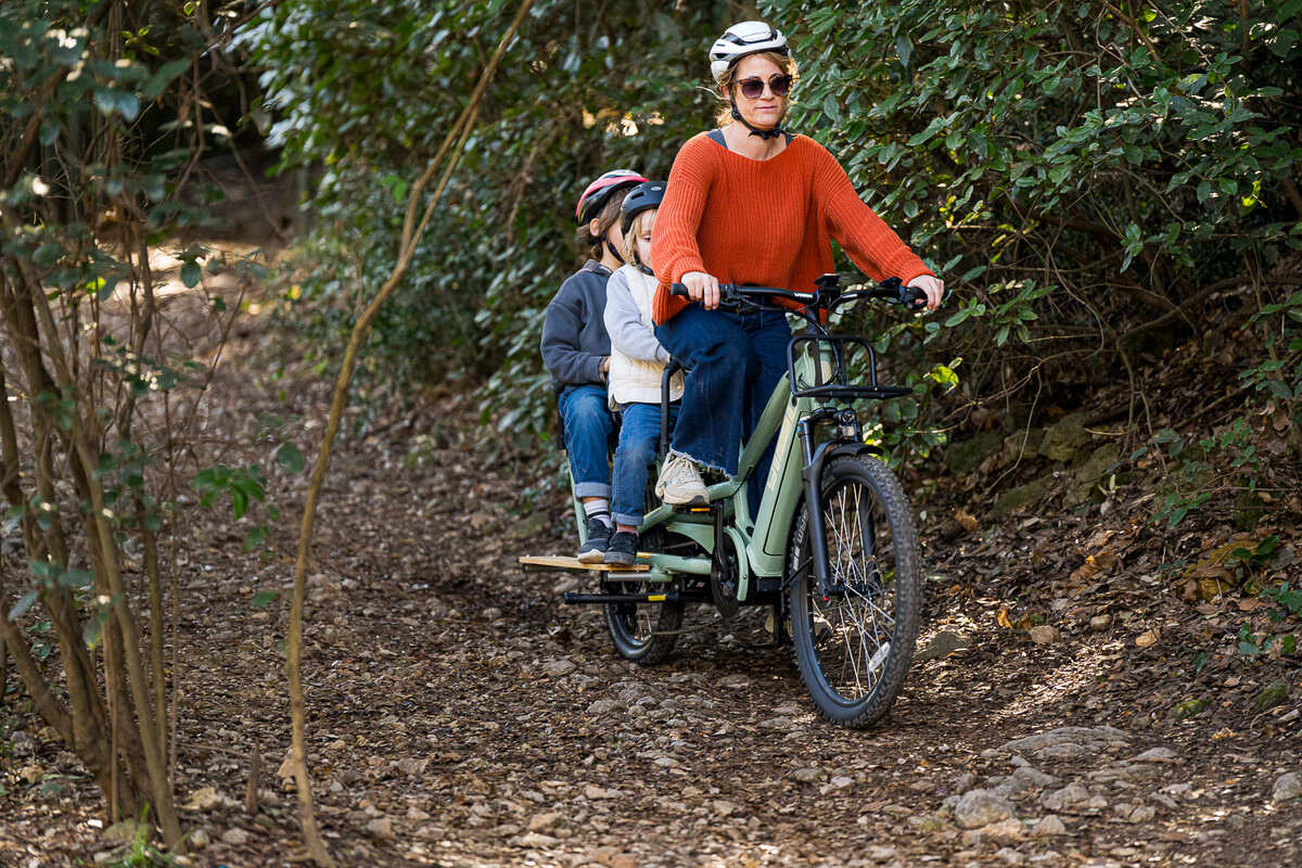 En person som bär en röd tröja och hjälm cyklar på en Yuba Spicy Curry Plus elektrisk lastcykel på en skogsstig, tillsammans med två hjälmförsedda barn. De navigerar smidigt genom tätt grönt löv och nedfallna löv och njuter av en äventyrlig familjeutflykt i naturen.