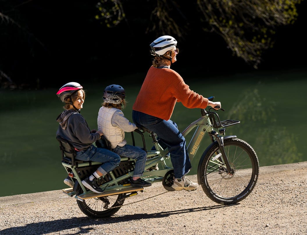 En person som bär en orange tröja cyklar på en Yuba Spicy Curry Plus longtail cykel längs en naturskön stig vid sjön, åtföljd av två barn som sitter säkert på den förlängda longtailen. Alla tre bär hjälmar när fläckigt solljus skiner genom de lummiga träden och reflekterar den skimrande sjön i bakgrunden.