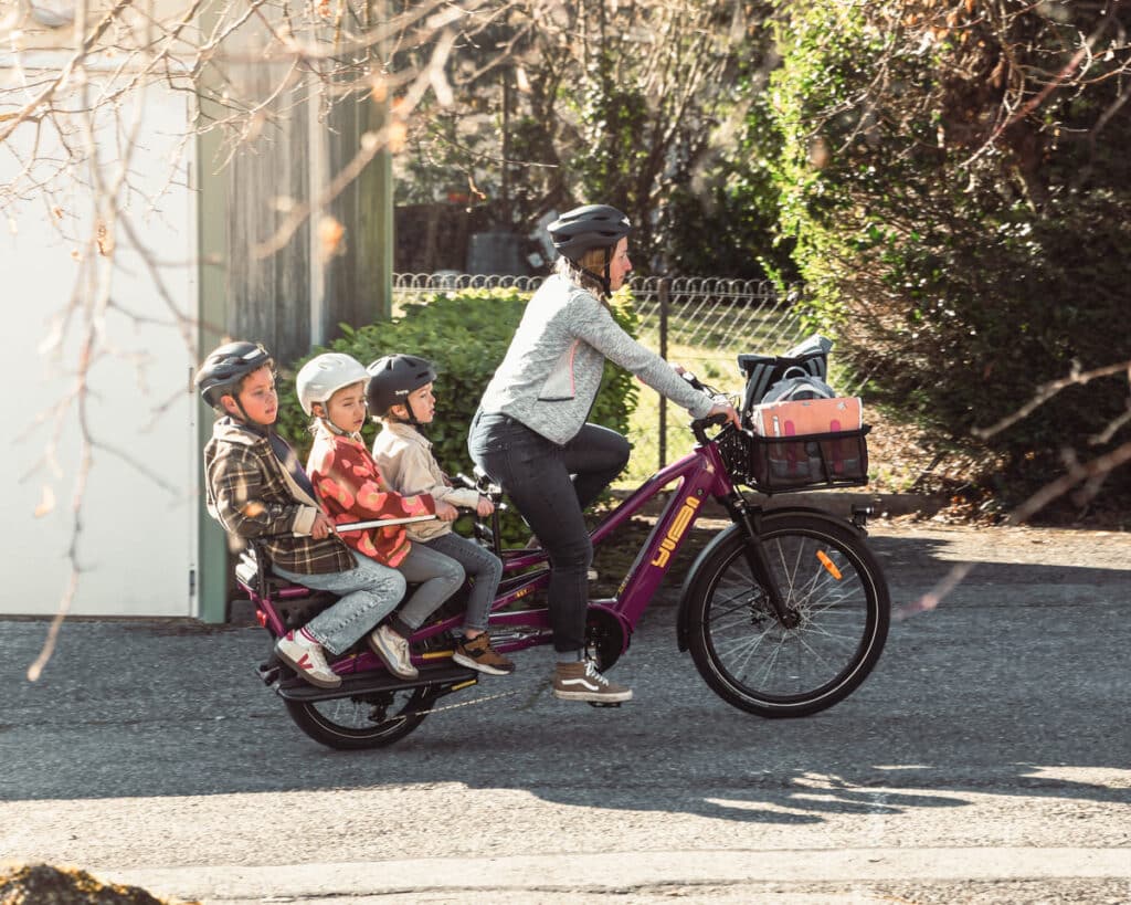 En vuxen med hjälm cyklar på en Yuba Spicy Curry Family Edition, en elastisk longtail-cykel i lila, med tre barn med hjälm sittande bakpå. Cykeln har en framkorg fylld med väskor medan de cyklar längs en solig bostadsgata.