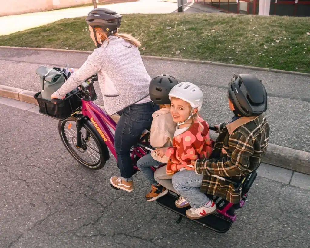 En vuxen cyklar på en Yuba Spicy Curry Family Edition-longtailcykel med tre barn på baksidan, och alla bär hjälm. Barnen sitter tätt ihop, och ett av barnen, som har en rödmönstrad jacka, ler mot kameran. Longtailcykeln med Bosch-motor står på en asfalterad gata nära en gräsyta.