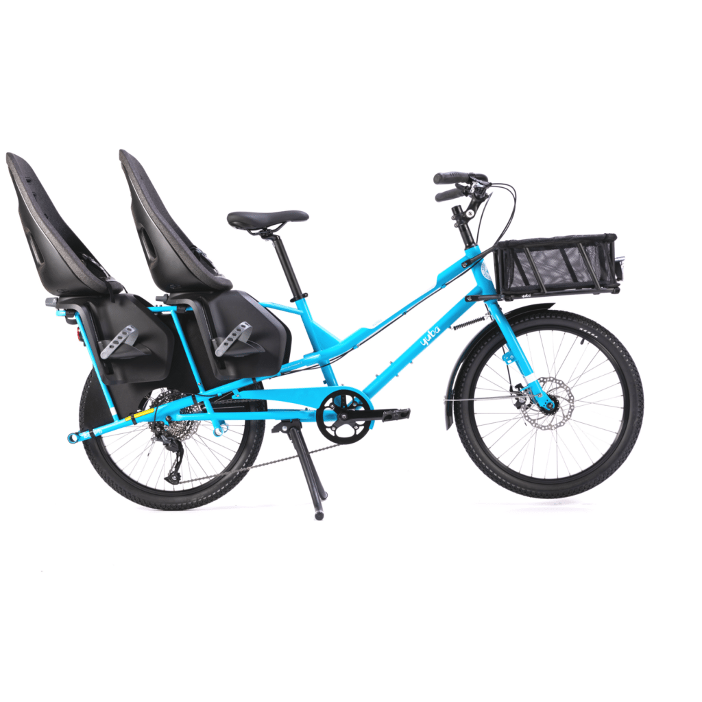 En livfullt blå Yuba Kombi-lastcykel av typen longtail visas mot en vit bakgrund. Den är utrustad med en robust metallkorg framtill, ett praktiskt mittstöd och två barnstolar monterade baktill för att transportera passagerare.