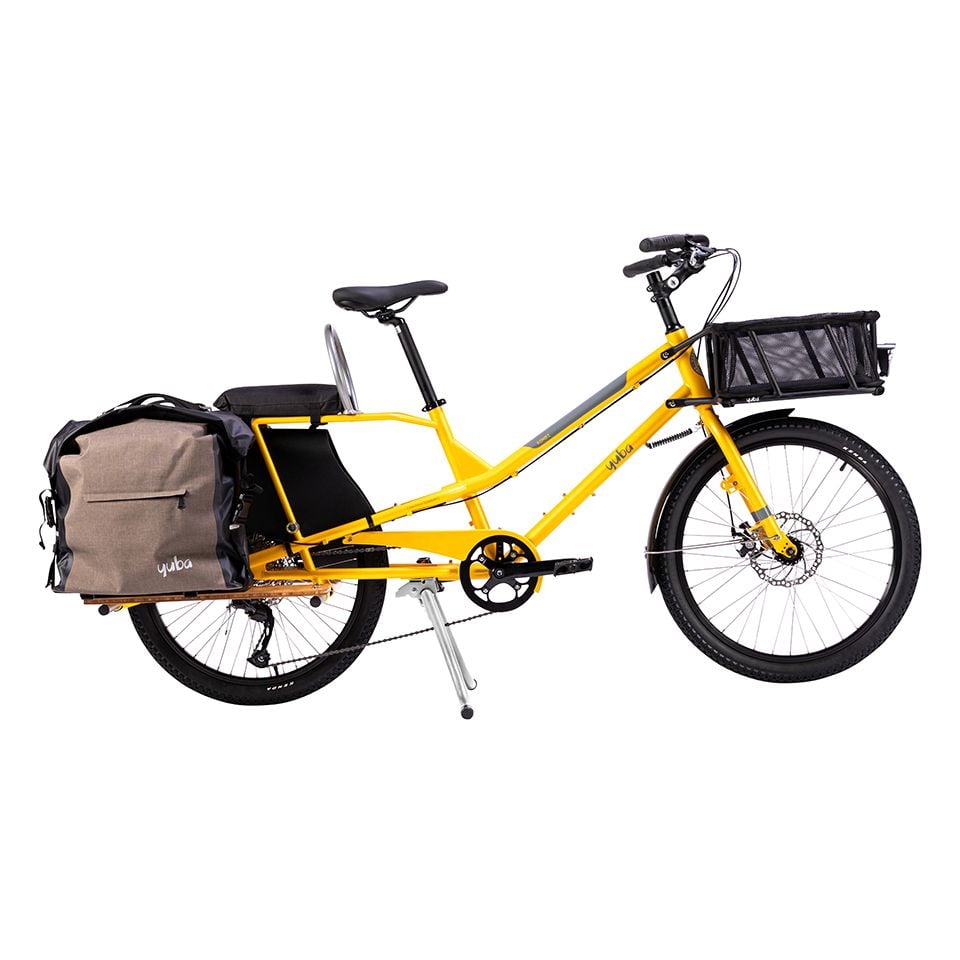 En gul Yuba Kombi-lastcykel (icke-elektrisk, longtail-modell) utrustad med en framkorg, en stor sidoväska på bakhjulet, två säten och en robust ram avsedd för extra last.