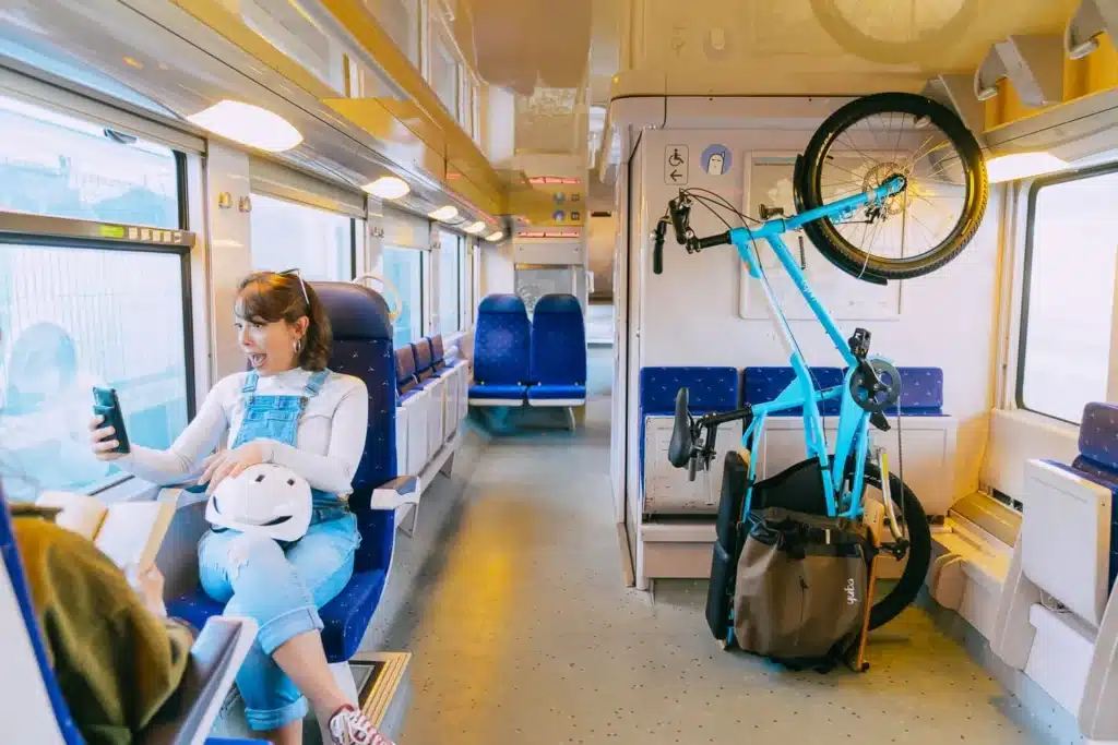 En glad kvinna i jeansoverall tar en selfie ombord på ett modernt, väl upplyst tåg. Hon håller en vit cykelhjälm i knät medan hennes blå Yuba Kombi lastcykel är säkert monterad på ett vertikalt cykelställ i närheten.