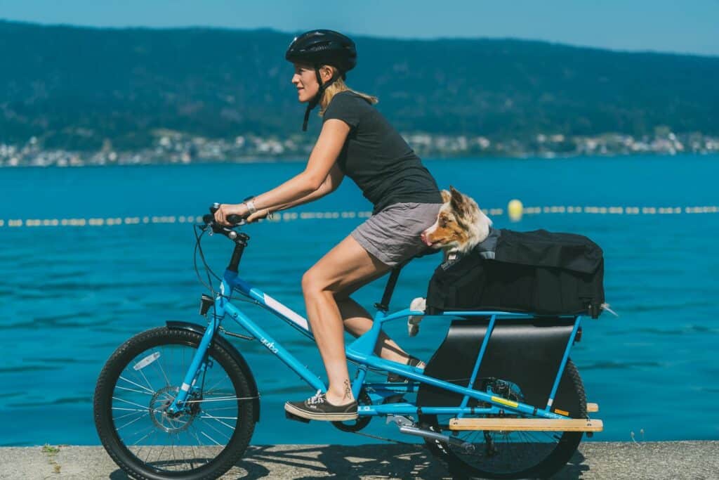 En person med hjälm cyklar på en blå Yuba Kombi vid en sjö, med en liten hund som sitter i en svart väska på bakre pakethållaren. I bakgrunden syns berg och vatten, vilket ger en vacker inblick i denna praktiska longtail-cykel.