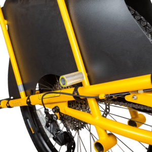 Närbild av bakdelen av en gul lastcykel mot svart bakgrund, som framhäver den robusta ramen, bakhjulet med synliga kugghjul och kedja, skyddsöverdrag och Kids Pegs – små fotstöd i metall monterade på ramen för att transportera passagerare.