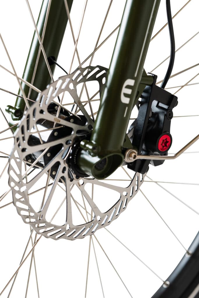 Närbild på framhjulet på Yuba Mundo Lux lastcykel, med en silverfärgad skivbromsrotor, ett svart bromsok med rött justeringsratt och en klargrön gaffel med synliga metallekrar.