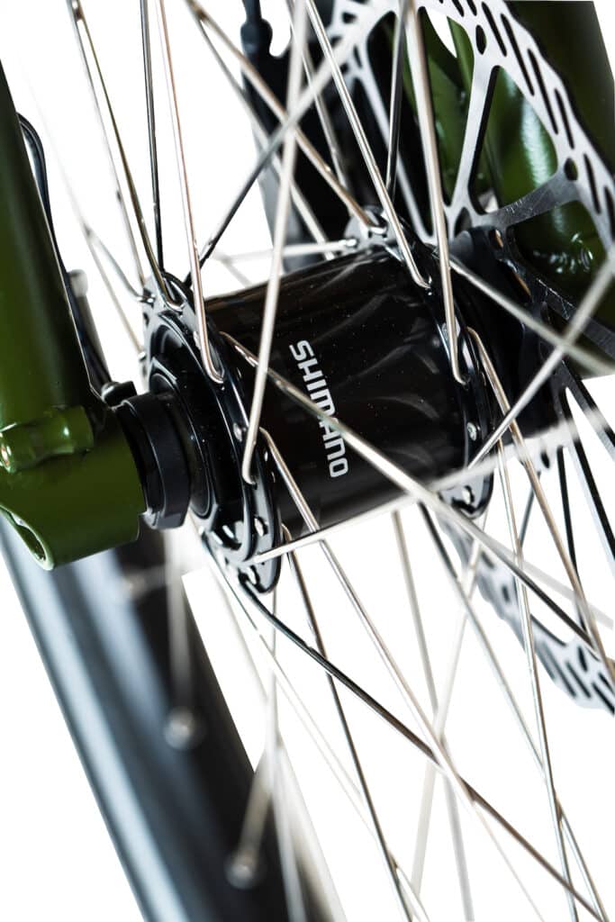 Närbild på ett Yuba Mundo Lux cykelframhjul med silverfärgade ekrar, ett svart Shimano-nav, skivbromsskiva och en grön framgaffel.