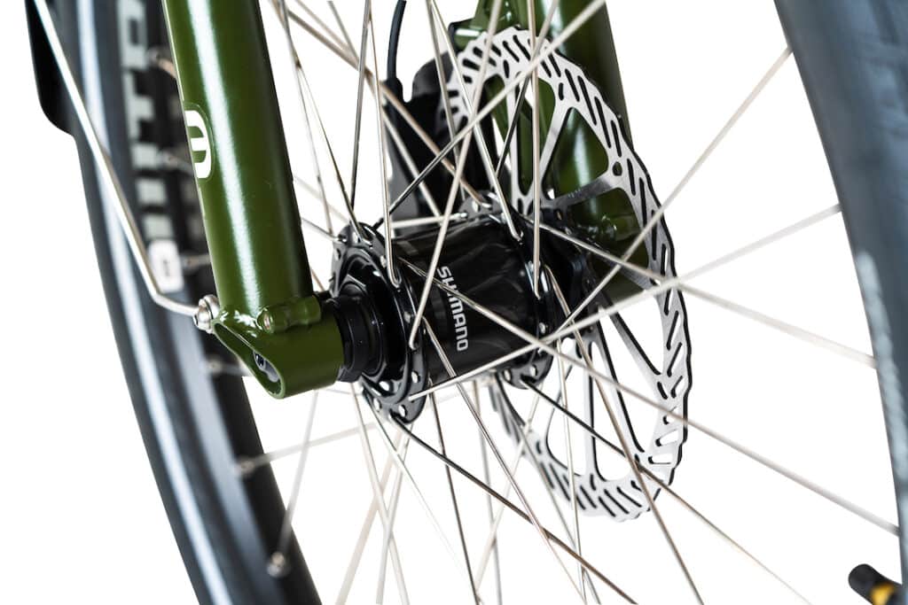 Närbild på ett framhjul på en Yuba Mundo Lux-lastcykel som visar silverfärgade ekrar i metall, Shimano-nav, skivbromsrotor och grön framgaffel mot en vit bakgrund.