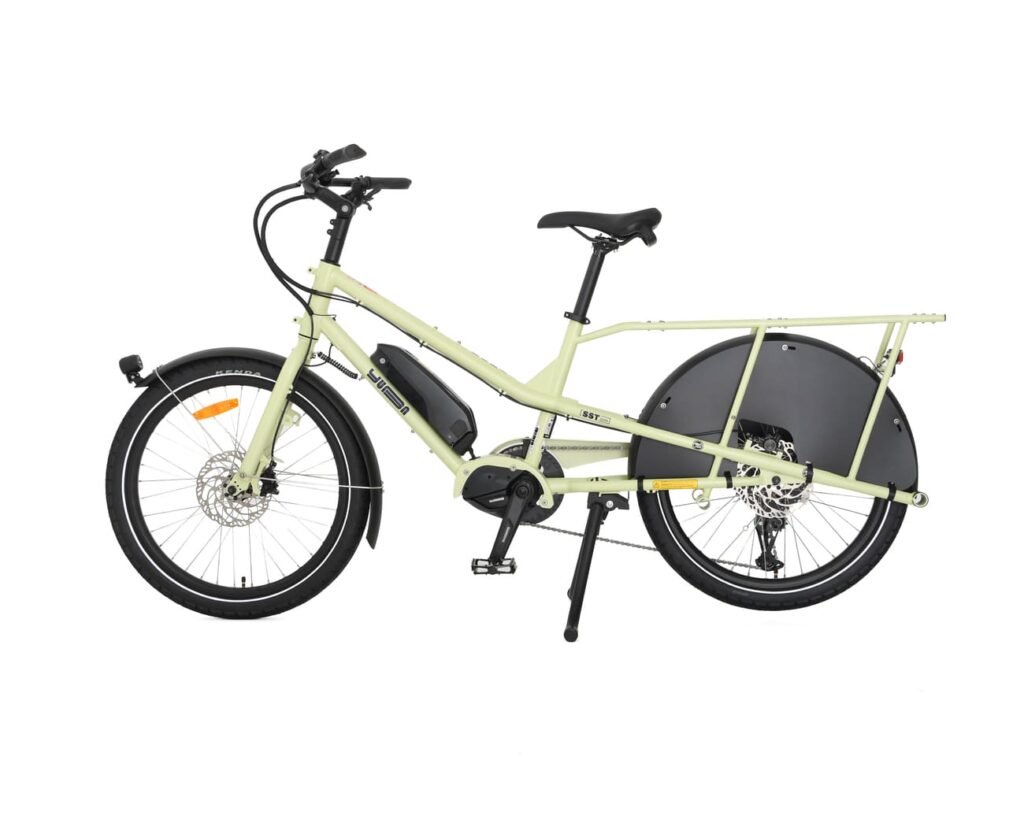 En elektrisk lastcykel av modellen Yuba Kombi EP5 i ljusgrönt, med svart bakhjulskåpa, mittmonterad motor, stänkskärmar fram och bak samt ett förlängt bakre pakethållare, avbildad i profil mot en vit bakgrund.