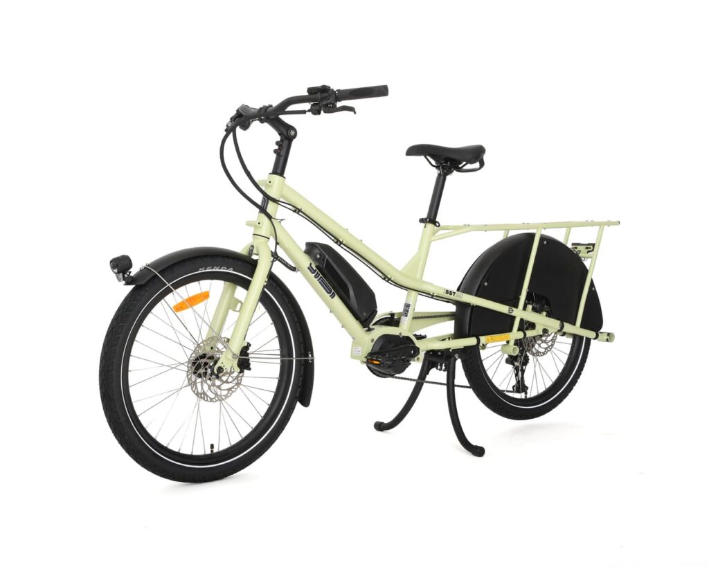 En ljusgul Yuba Kombi EP5, en eldriven lastcykel med ett långt bakre pakethållare, svarta stänkskärmar och en robust ram, visas i sidovy mot en vit bakgrund.
