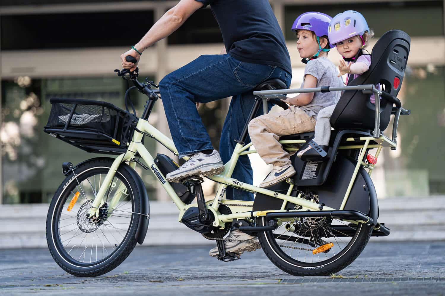 En vuxen cyklar på en Yuba Kombi EP5-lastcykel med två barn med hjälm på baksätet. Cykeln har en korg framtill, och gruppen verkar cykla på en asfalterad väg under sin provtur.