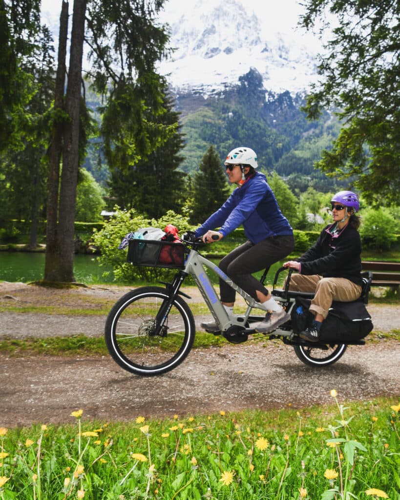 Två cyklister med hjälm cyklar längs en parkväg på en Spicy Curry x POW EZ Family Edition (2026) med korg, sidoväskor och vildblommor; träd och berg utgör den vackra bakgrunden.